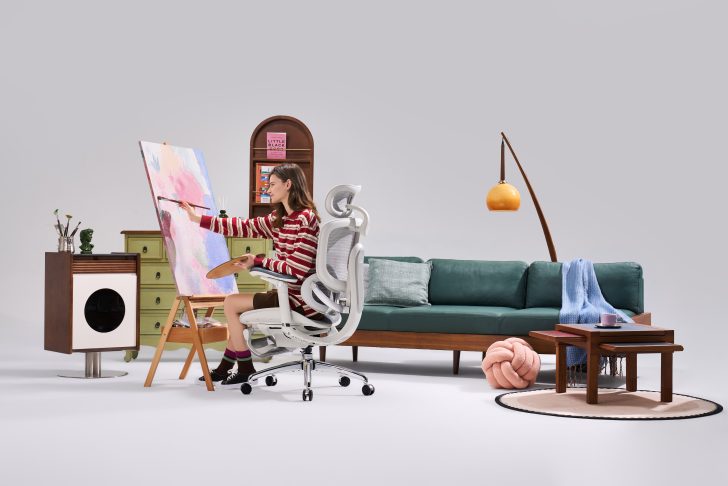 A person sitting in a modern white office chair is painting on a large canvas with various home decor items in the background.