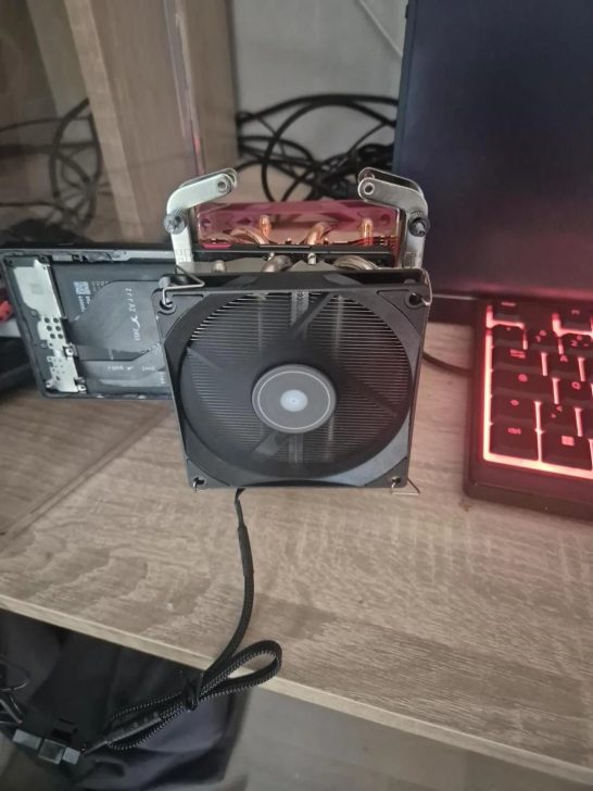A desktop cooling fan is humorously mounted on a laptop keyboard, with visible wiring and a backlit red keyboard to the side.