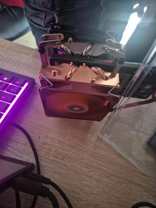 A large CPU cooler with exposed heat pipes is mounted on a computer processor near a mechanical keyboard with purple backlighting.