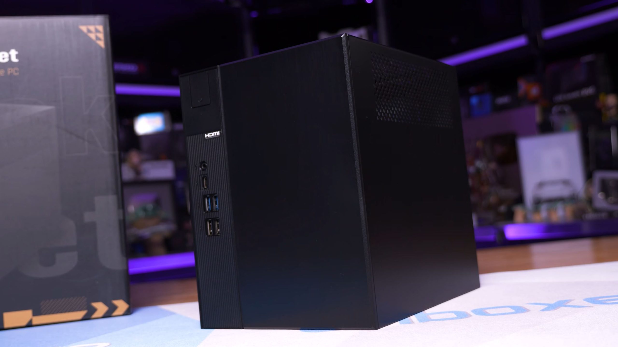 A black, compact case form-factor PC is placed beside its packaging box, with visible HDMI, USB, and audio ports on its front panel.