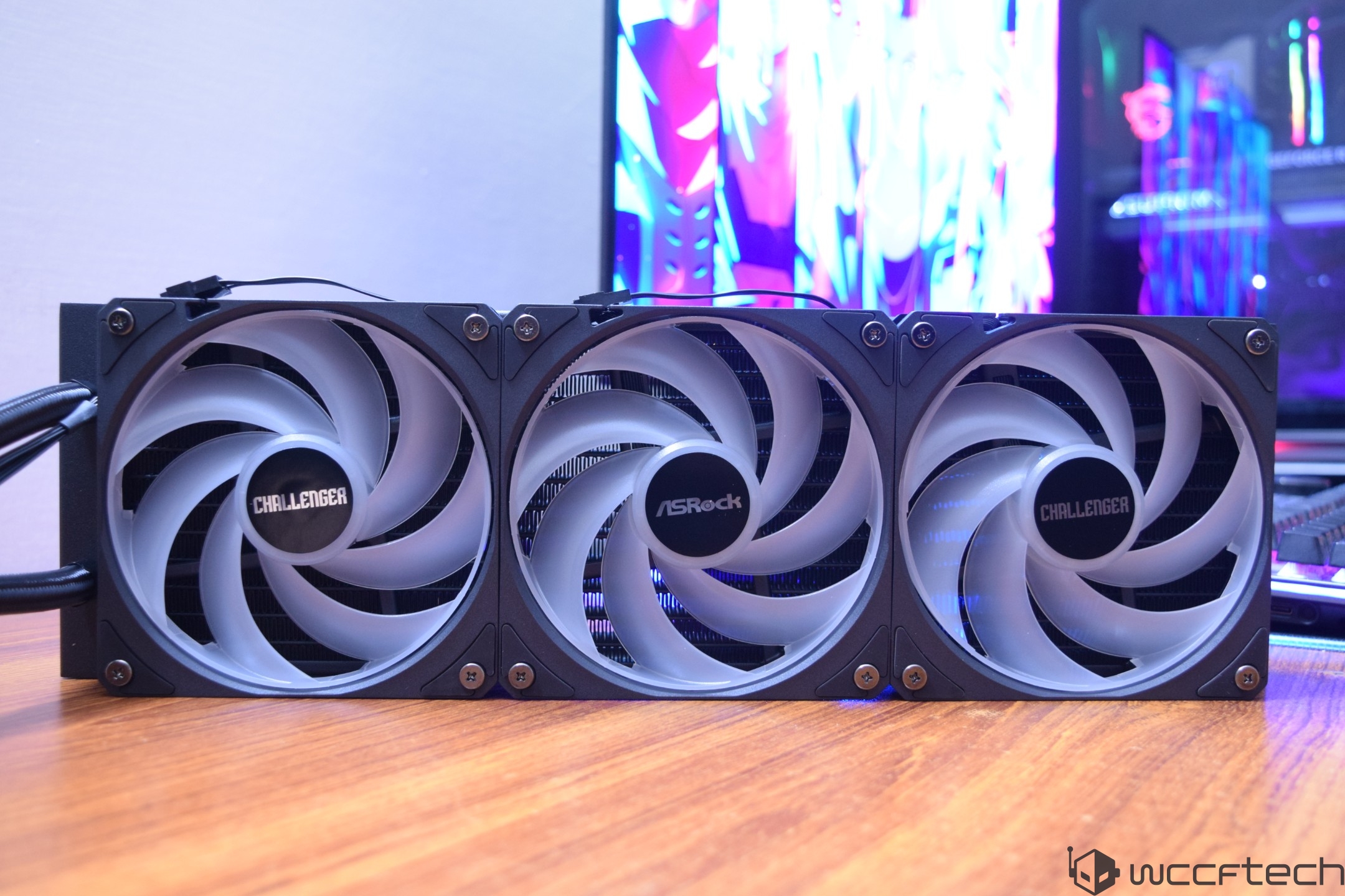 An ASRock cooling system with three large fans labeled 'CHALLENGER' is displayed on a wooden surface with a colorful screen in the background.