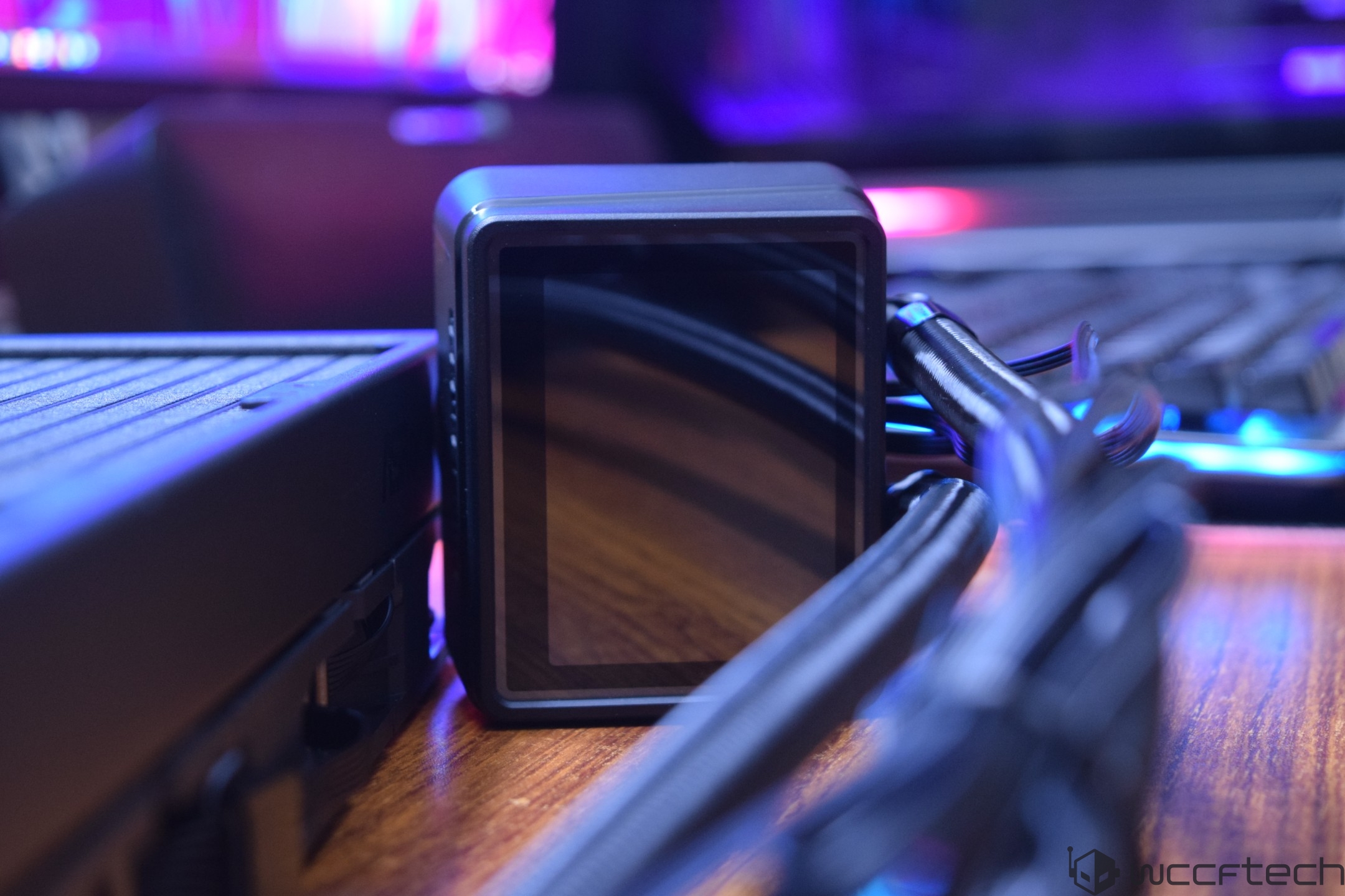 A close-up of a black, square display unit on a computer cooling system with visible braided cables in a tech setup.