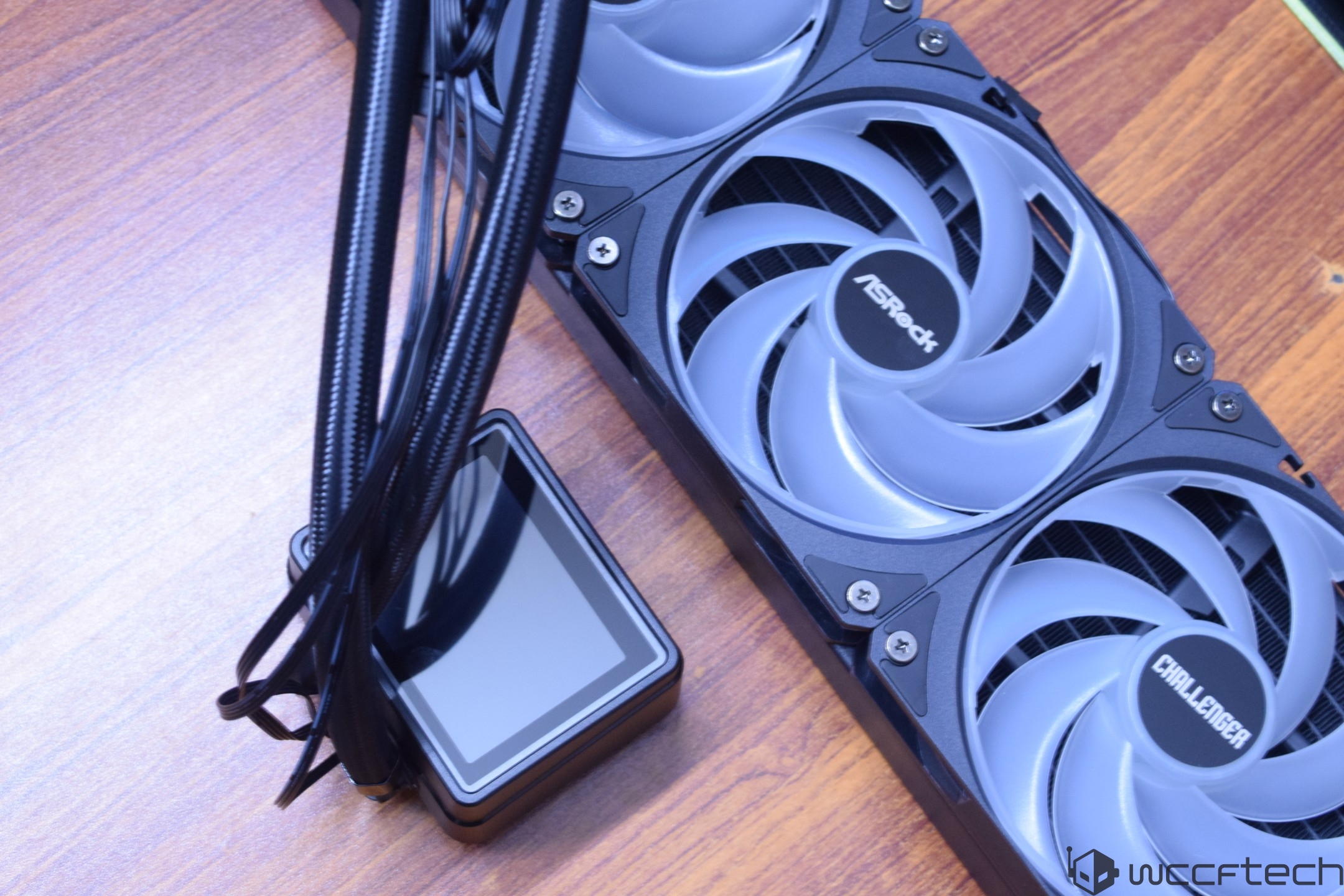 An ASRock Challenger liquid cooling system with visible fan blades and a braided cable is placed on a wooden surface.