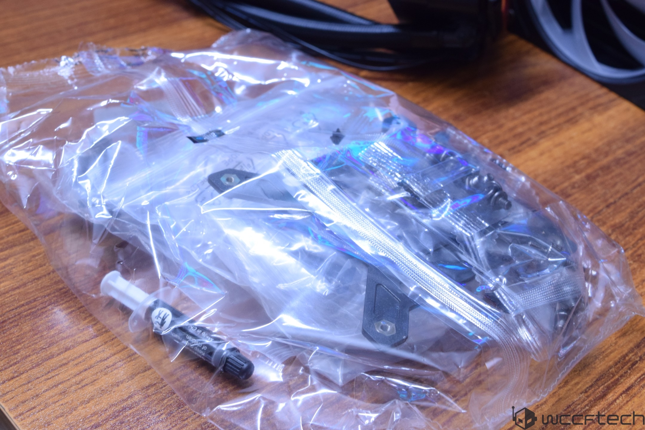A plastic bag containing computer hardware components and a small syringe of thermal paste on a wooden surface, with Wccftech branding in the corner.