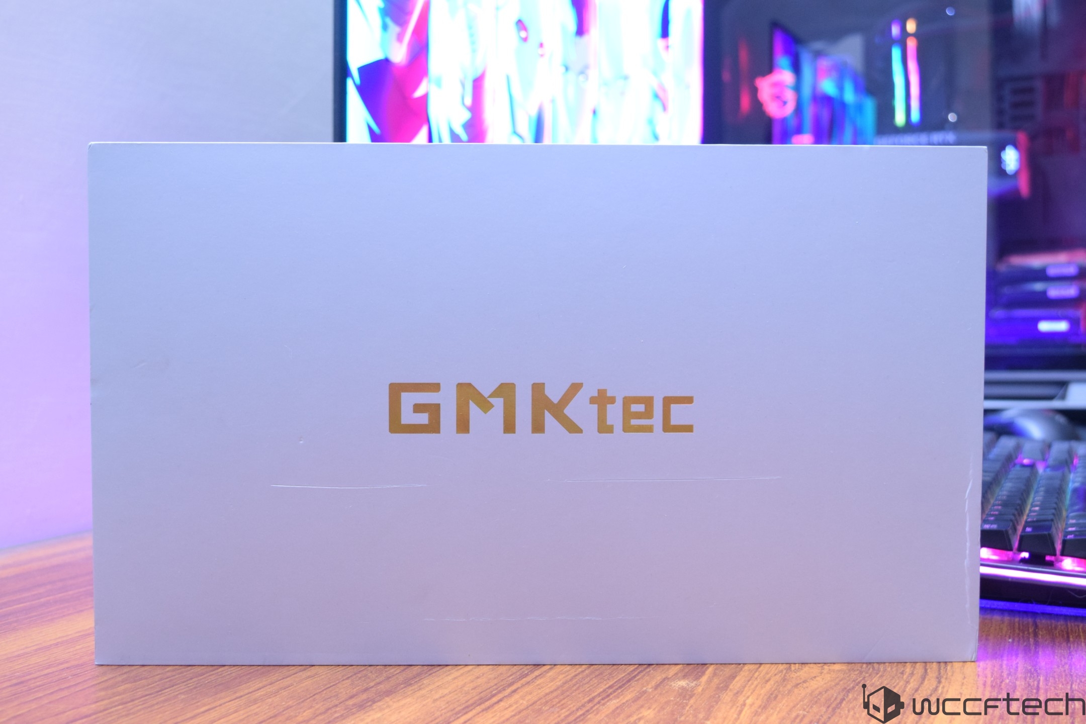 A box labeled 'GMKtec' is placed on a wooden desk in front of a computer setup with colorful LED lights in the background.