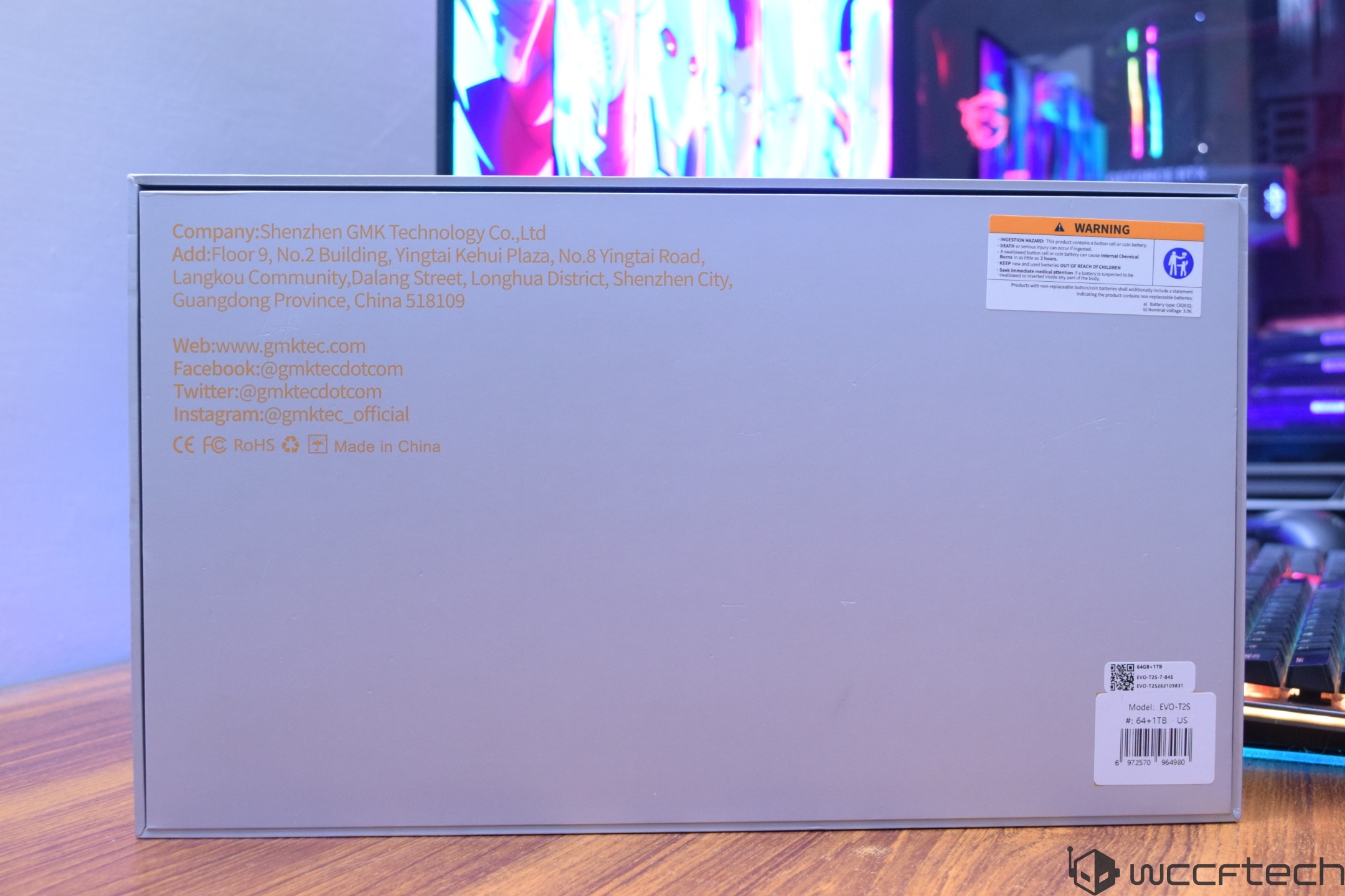 The back of a GMK Technology product box displays the company's contact information, a warning label, and model number 'EVO-TK5,' with colorful computer monitors in the background.
