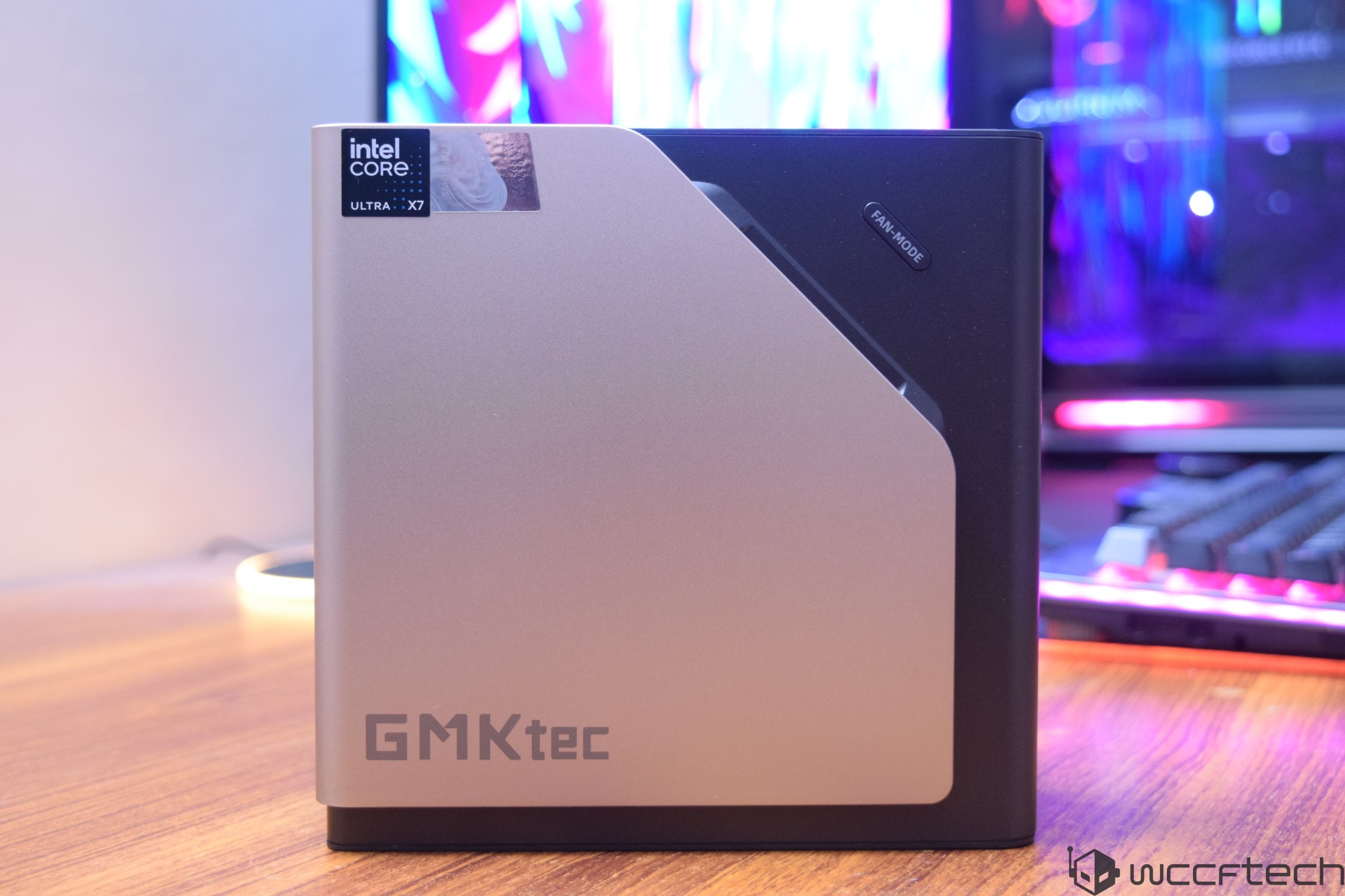 A GMKtec mini PC features an 'Intel Core Ultra X7' label and a 'FAN MODE' button on a desk with a colorful gaming setup in the background.
