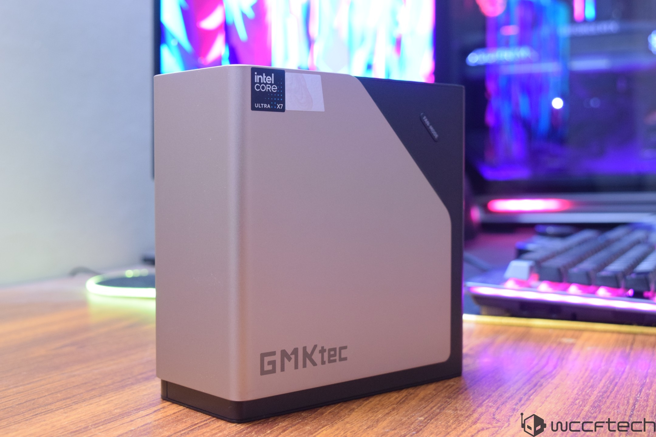 A GMKtec mini PC with an 'Intel Core Ultra X7' sticker is positioned on a wooden desk in front of a brightly lit gaming setup.