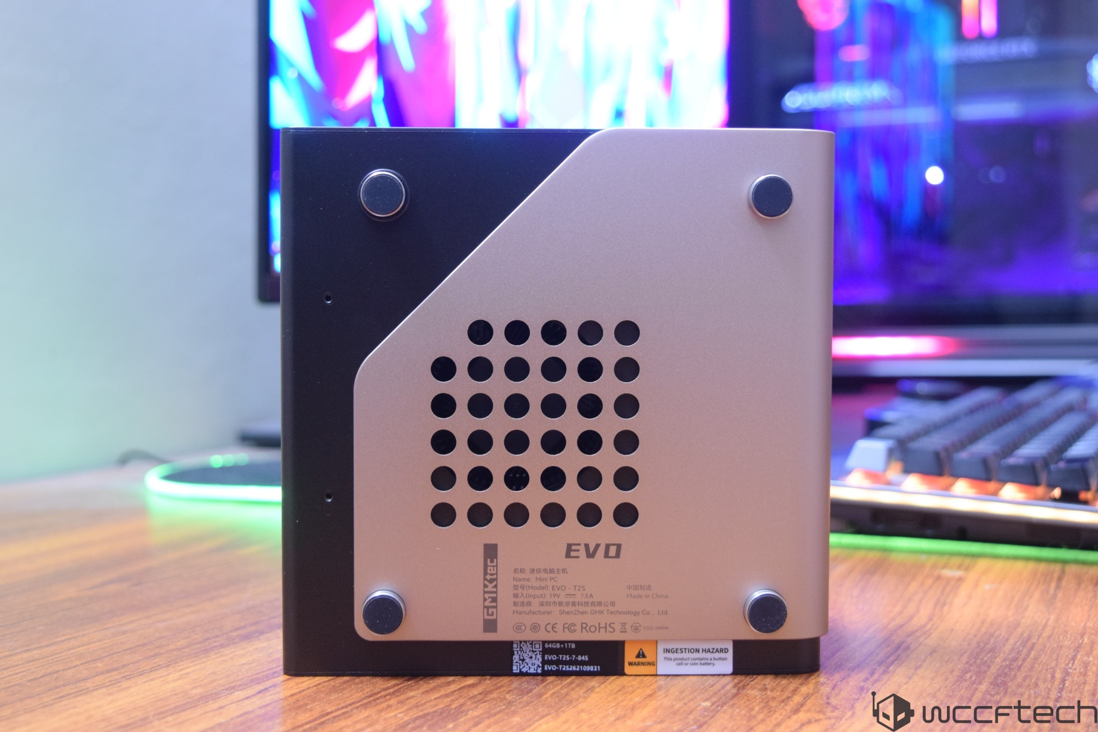 The back panel of a GMKtec Mini PC EVO model T8 is shown on a wooden desk with a colorful monitor and keyboard in the background.