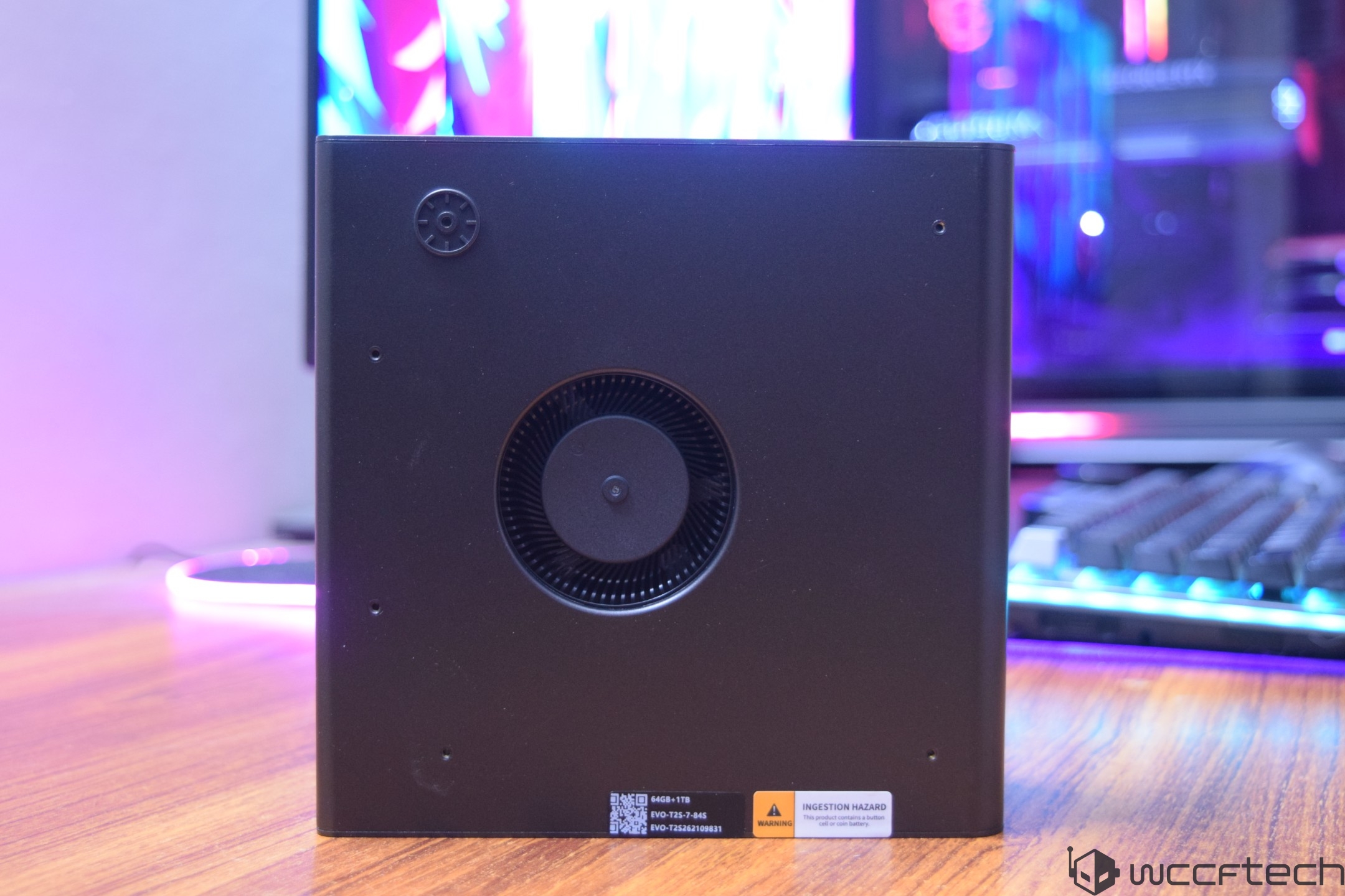 A black cube with a visible ventilation fan and a label reading 'INGESTION HAZARD' is placed on a wooden desk in front of a gaming setup with RGB lighting.