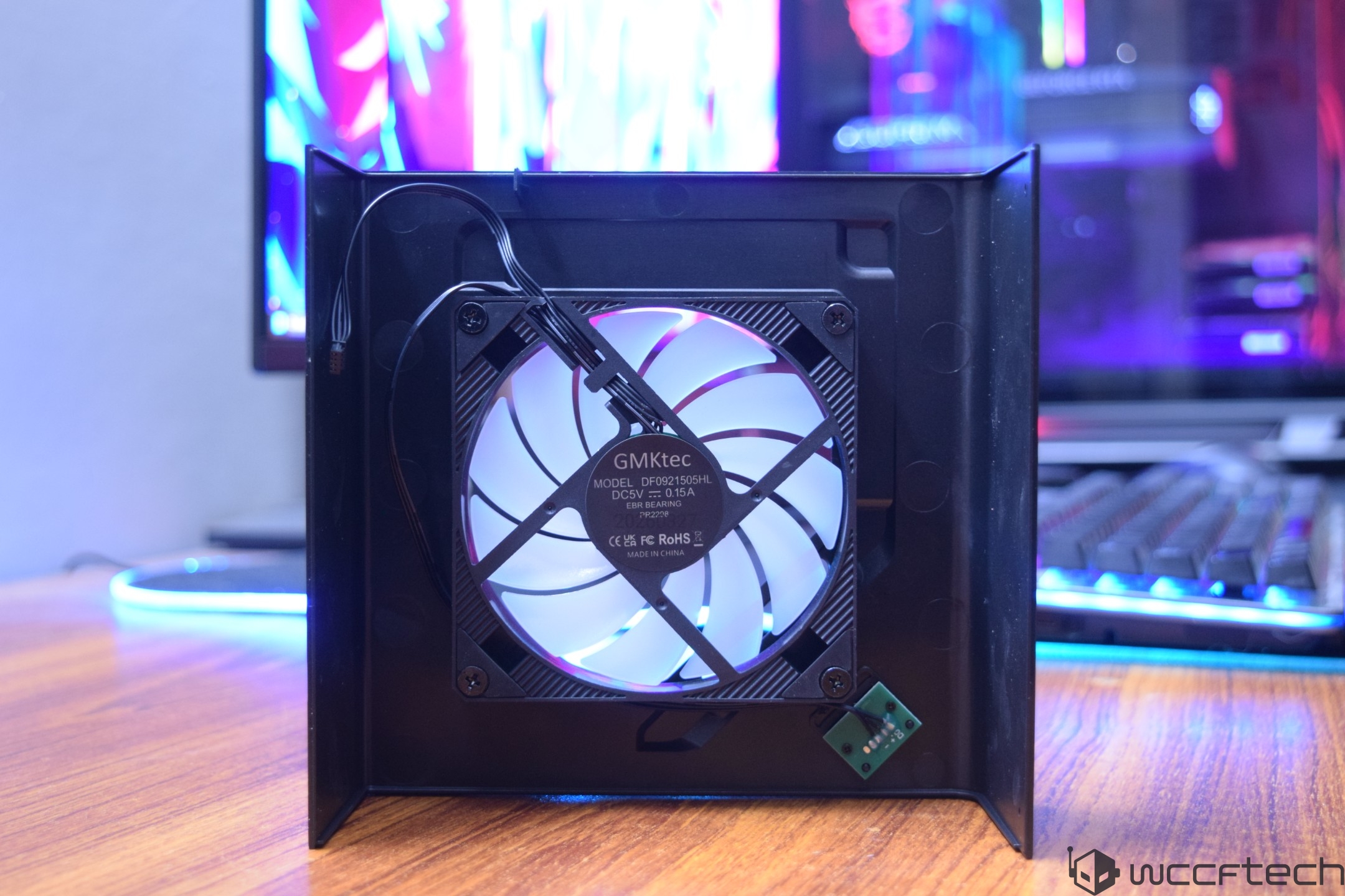 A GMKtec fan model DF09215Q5HL with visible 'DC5V 0.15A' text is mounted inside a black chassis, placed in front of a colorful computer monitor.