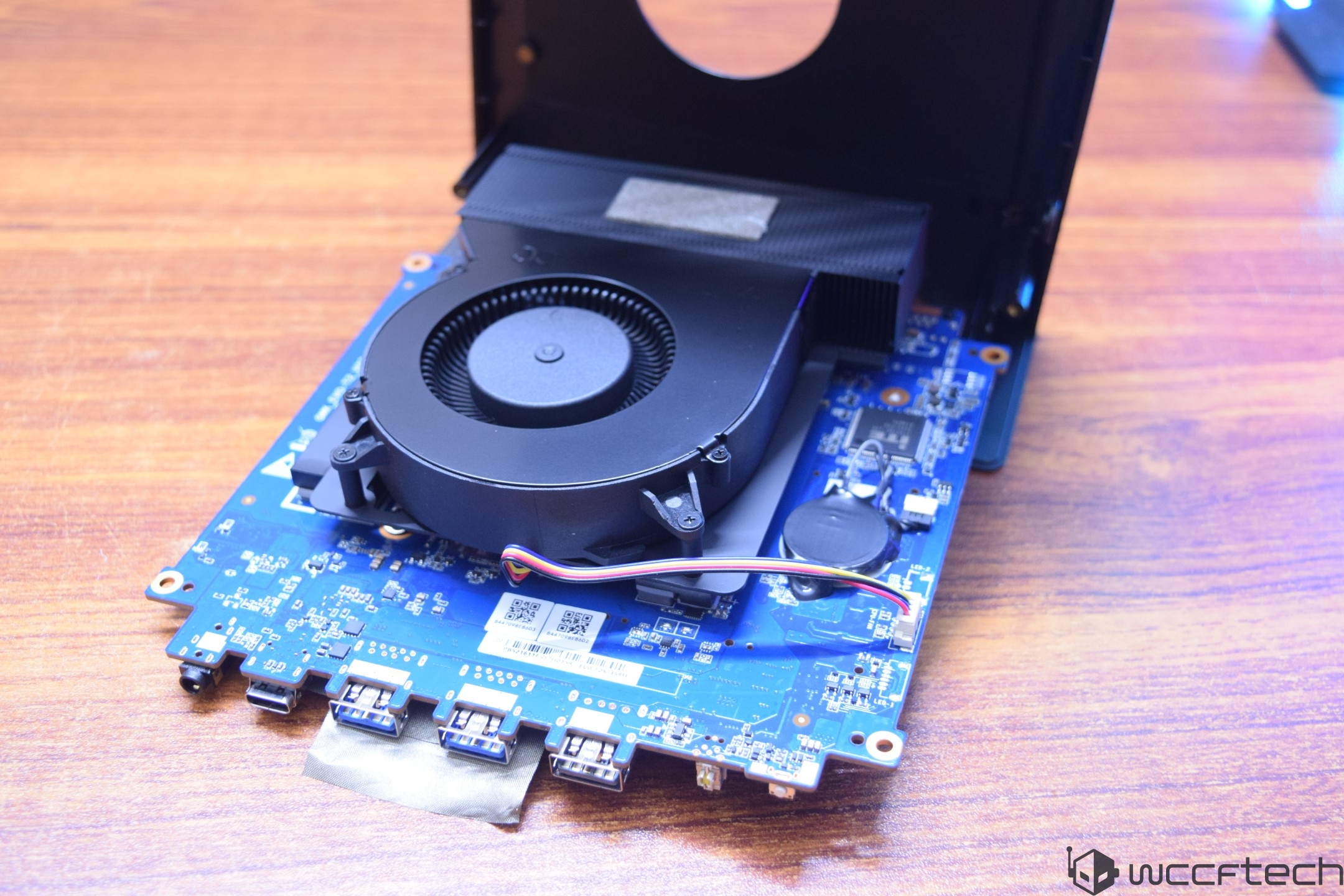 A small form factor desktop motherboard with a black cooling fan and visible USB ports is displayed on a wooden surface.