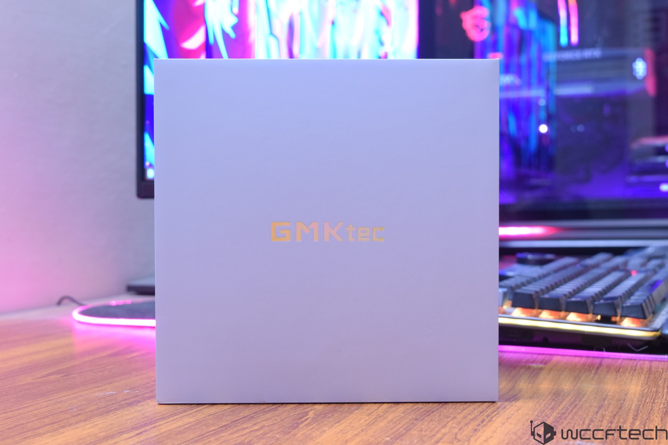 A GMKtec product box is placed on a wooden desk in front of a gaming setup with a colorful, illuminated background.