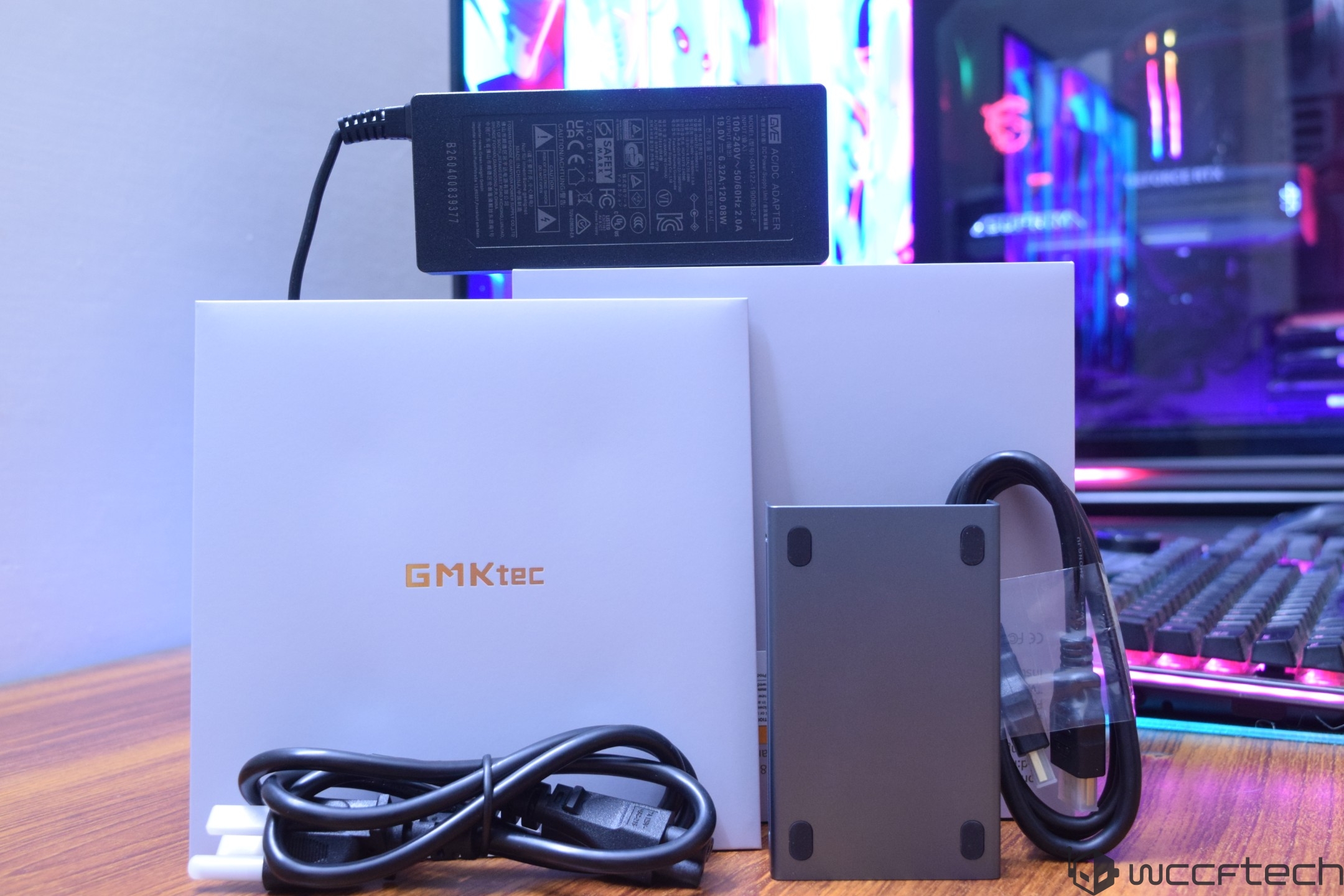 A GMKtec product box with branding, alongside a power adapter, cables, and a metallic peripheral, is displayed on a wooden surface with a colorful PC setup in the background.