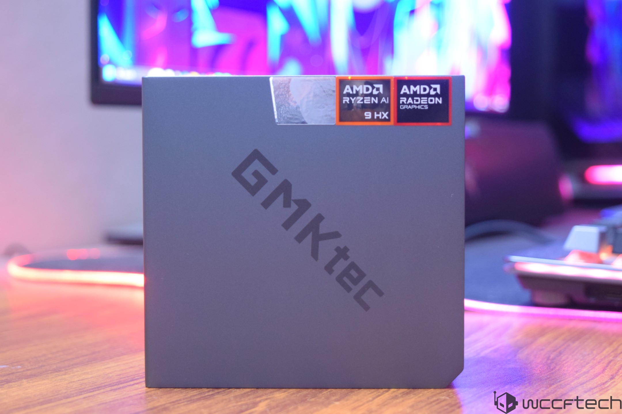 A GMKtec device box displays logos for 'AMD Ryzen AI 9 HX' and 'AMD Radeon Graphics' on a desk with a blurred screen in the background.