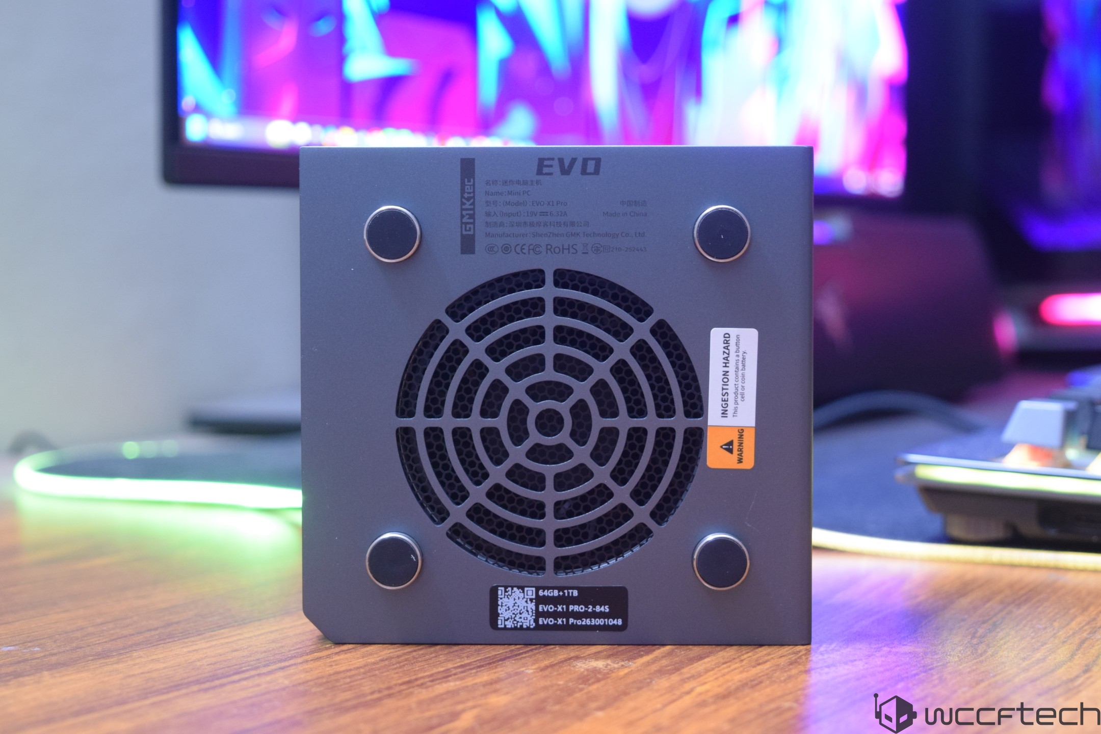 A GEEKOM Mini IT8 with visible '64GB+1TB,' 'EVO-X1 PRO-2-8435,' and 'EVO-X1 Pro26300104B' labels is on a desk, with a monitor displaying a colorful abstract background in the distance.