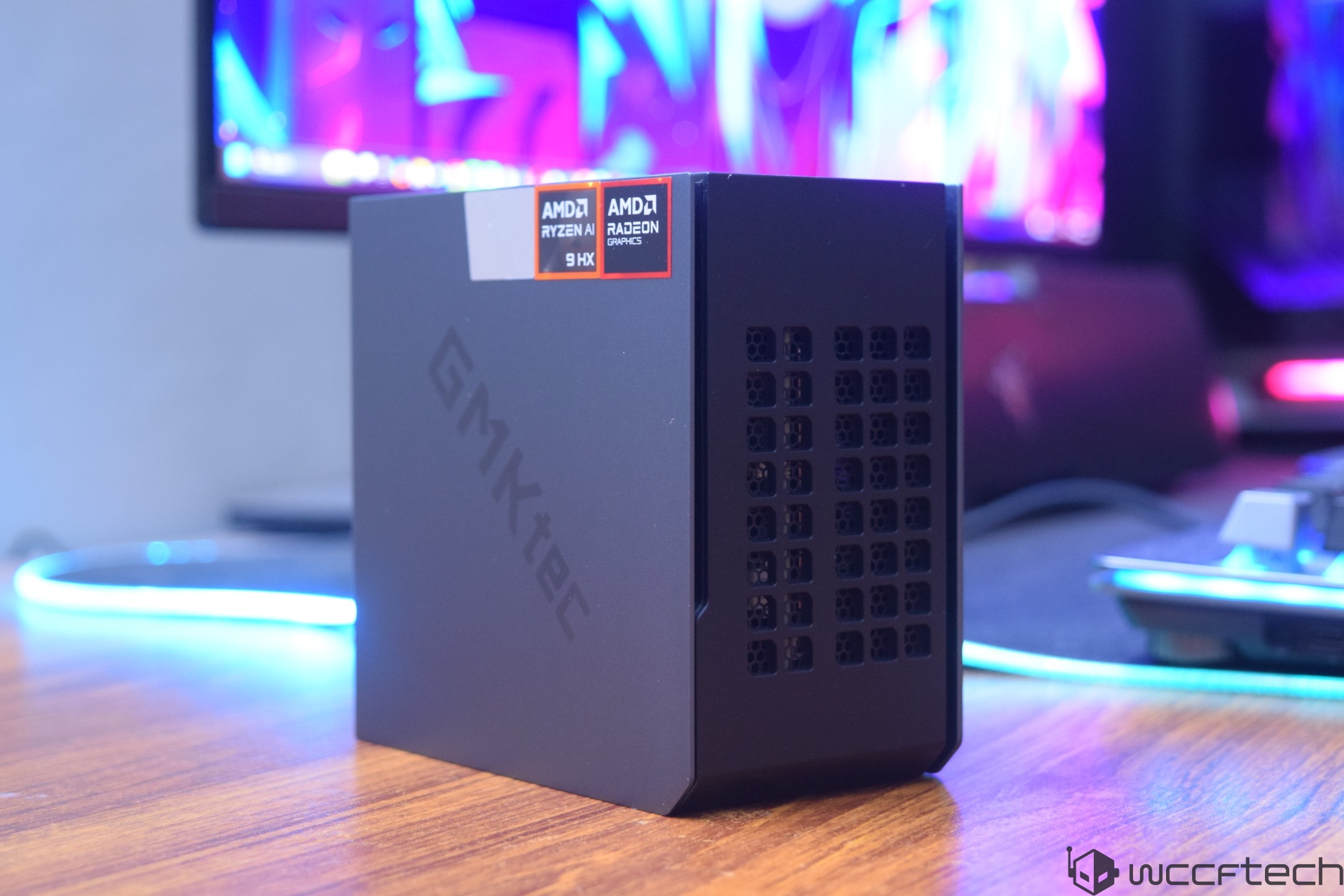 A GMKtec mini PC with AMD Ryzen 9 HX and Radeon graphics logos is displayed on a wooden desk with a colorful monitor in the background.
