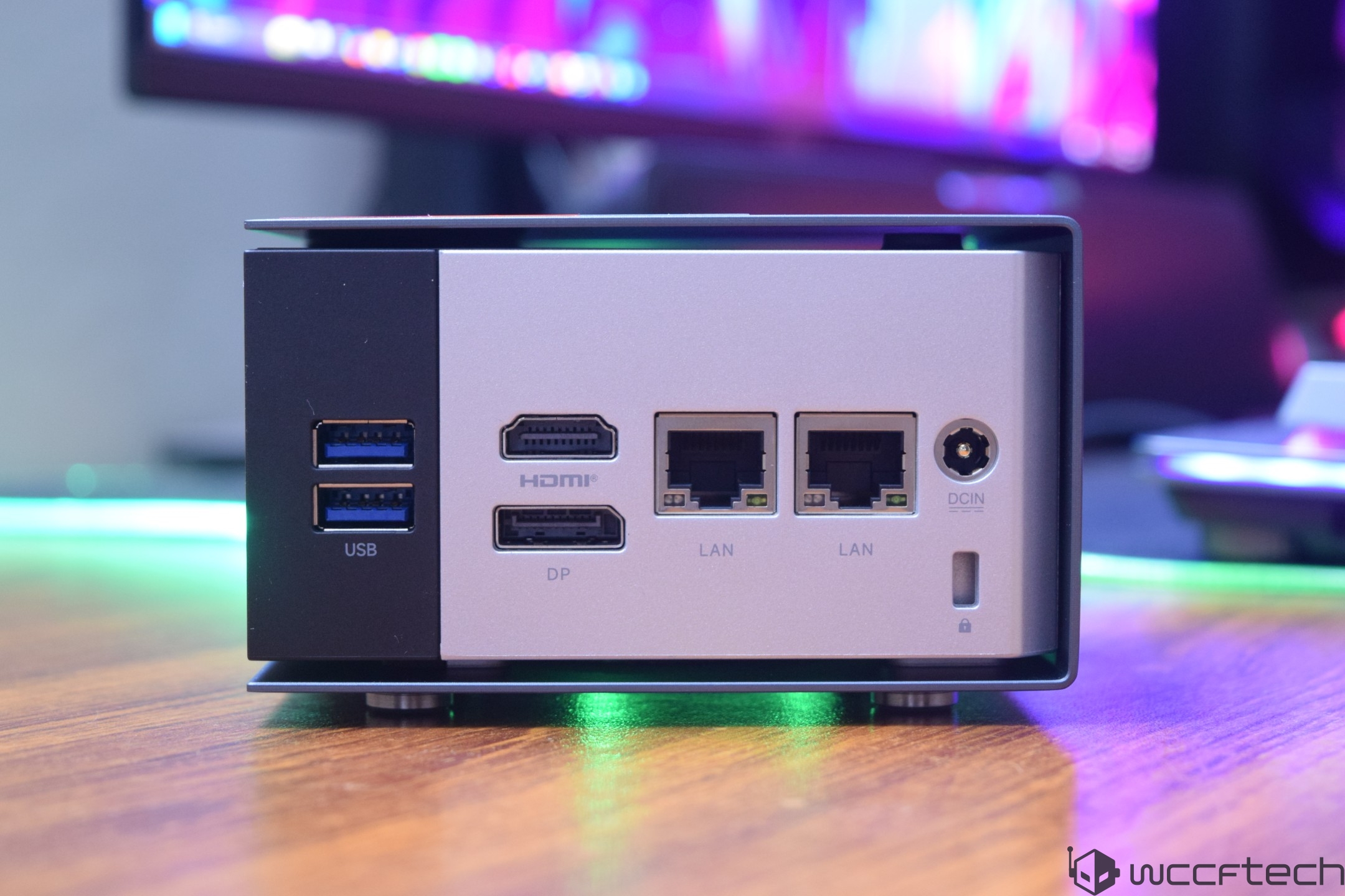 The back panel of a mini PC showing two USB ports, HDMI, DP, two LAN ports, and a DCIN connector on a wooden surface with a blurred colorful background.