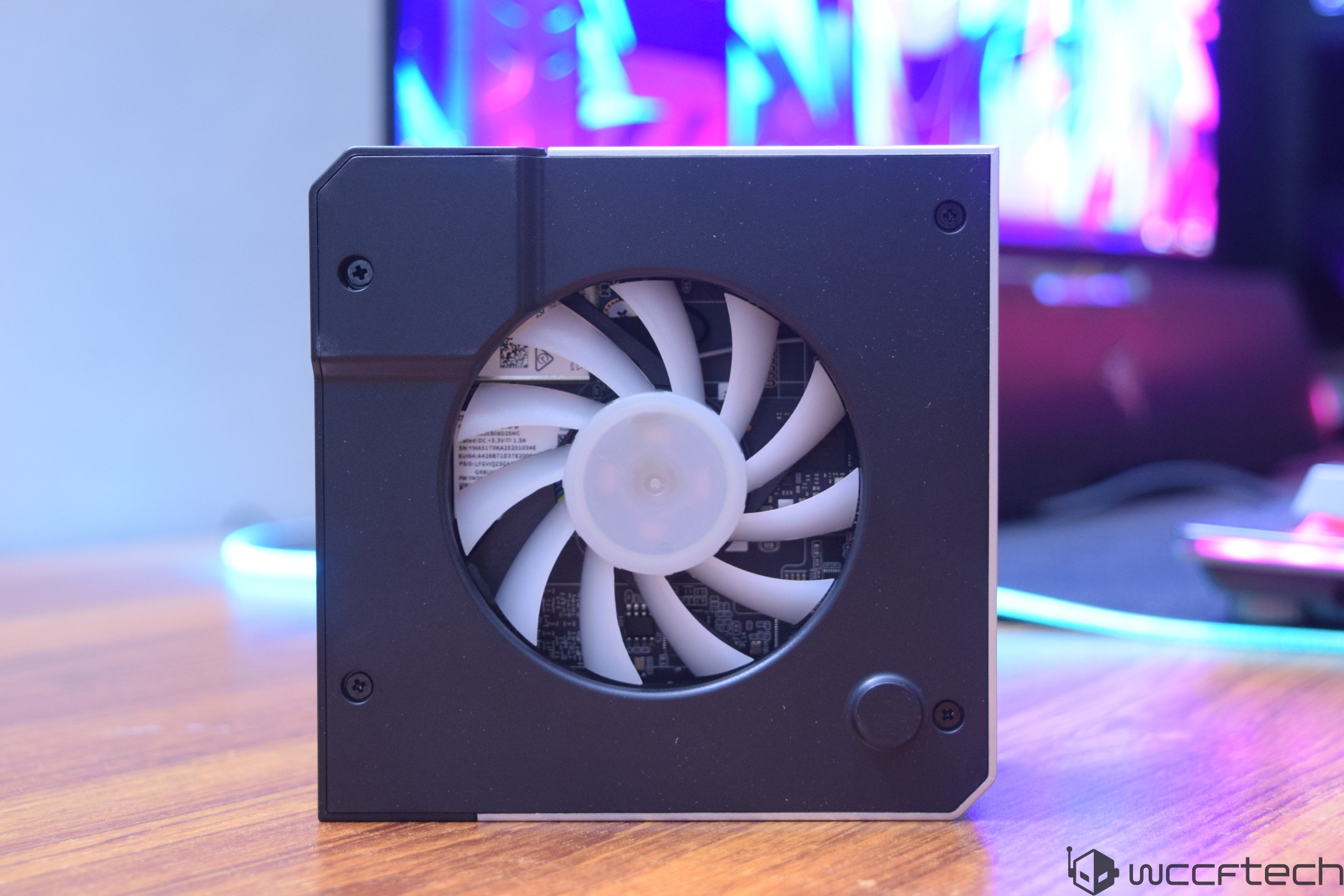 A close-up of a small, unbranded cooling fan unit on a wooden surface with a colorful monitor background.
