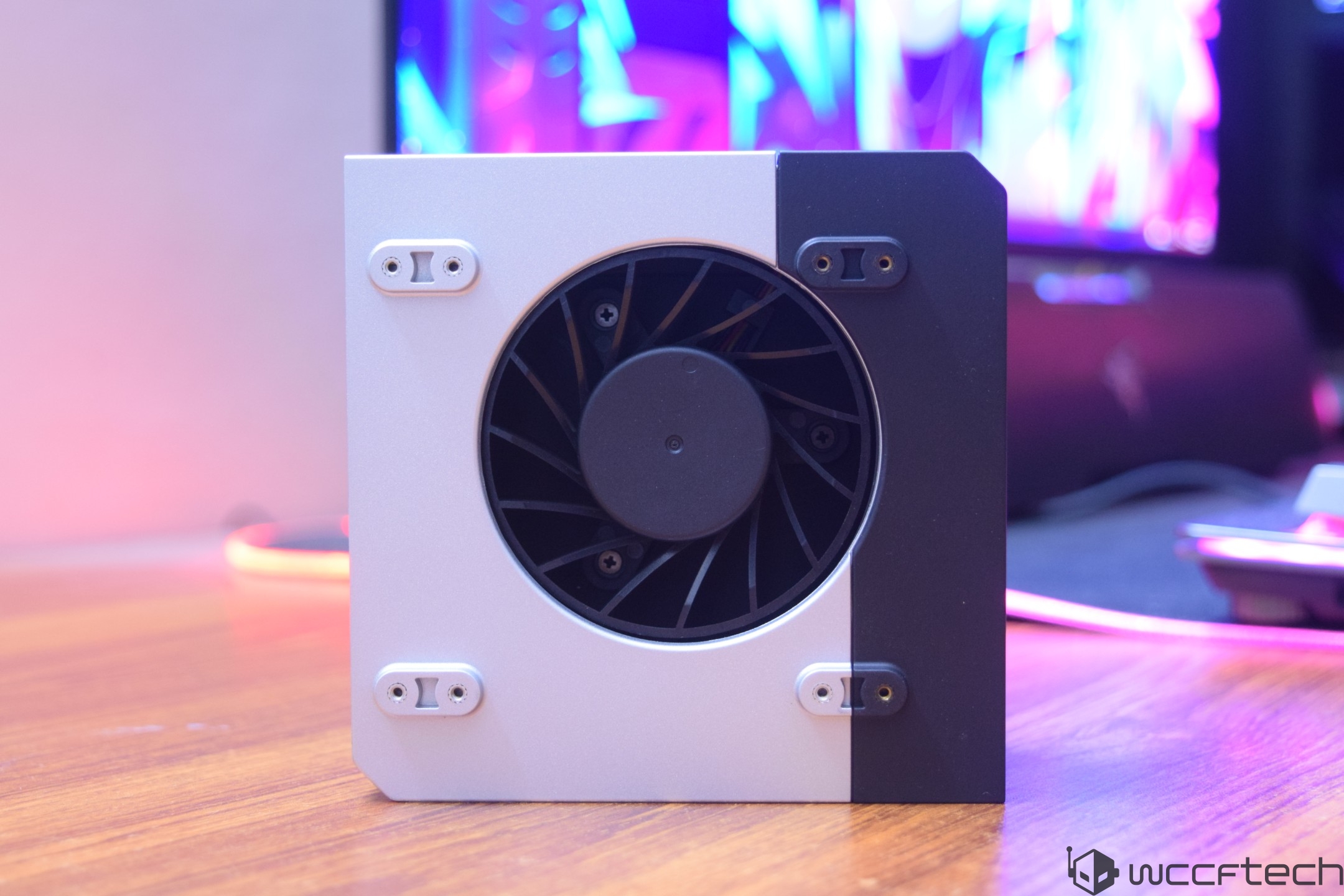 A close-up of a small desktop cooling fan with a circular fan design, set against a two-tone, white and black casing on a wooden surface, with a soft-focus computer setup in the background.