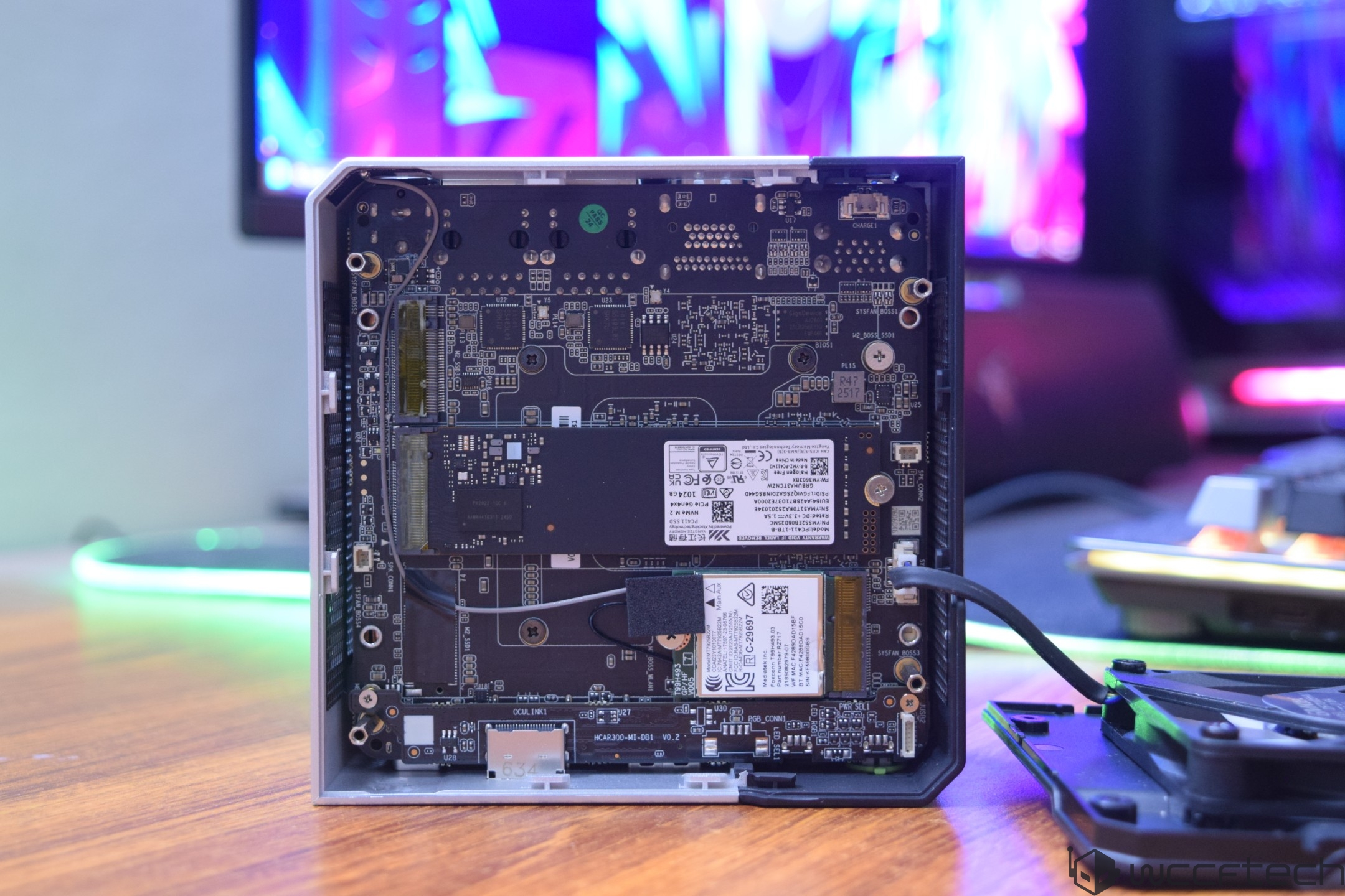 The interior of an unbranded mini PC case showing the motherboard with a Kingston M.2 NVMe SSD and a Wi-Fi card installed on a wooden desk, with blurred RGB-lit screens in the background.
