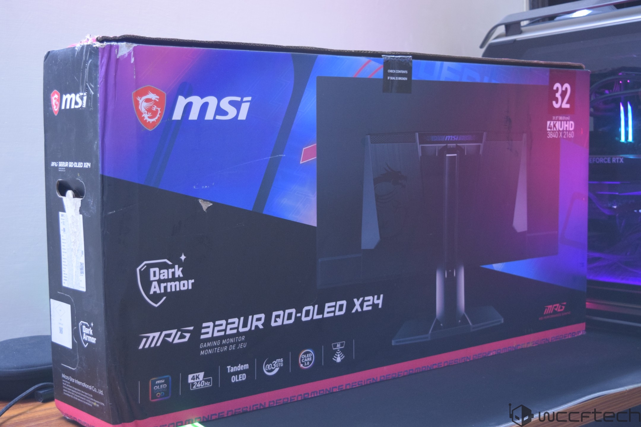 The packaging for an MSI MAG 322UR QD-OLED X24 gaming monitor is displayed next to a gaming PC.