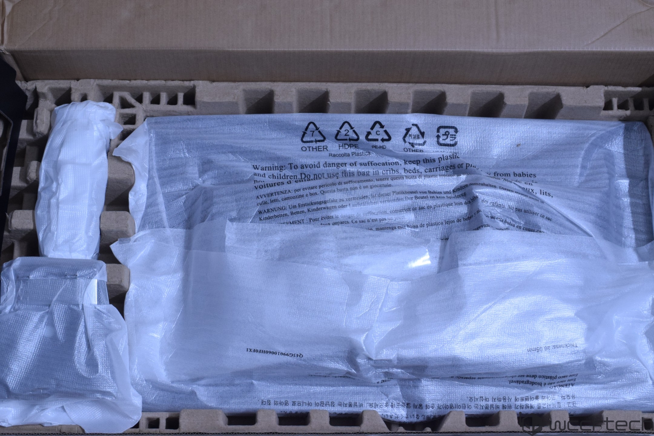 A large unbranded monitor is securely packaged in a cardboard box with protective foam inserts and covered with plastic featuring safety warnings in multiple languages.