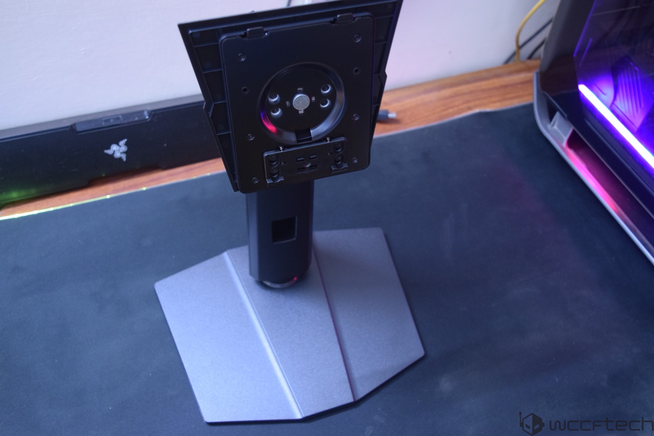 The image shows a monitor stand without a screen, positioned in front of a Razer keyboard and next to a gaming PC with a glowing purple interior.