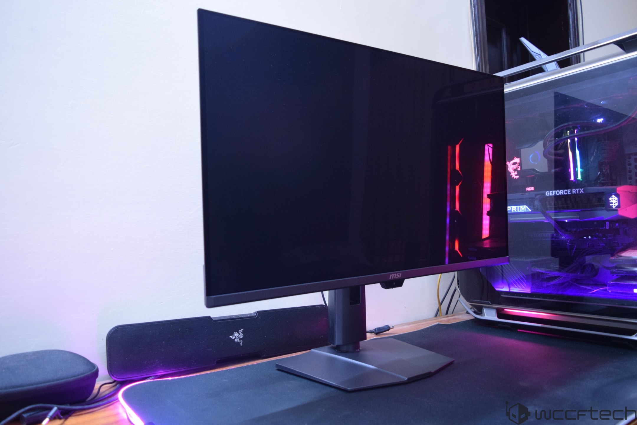An MSI monitor is positioned on a desk beside a transparent gaming PC case featuring GeForce RTX branding and RGB lighting.