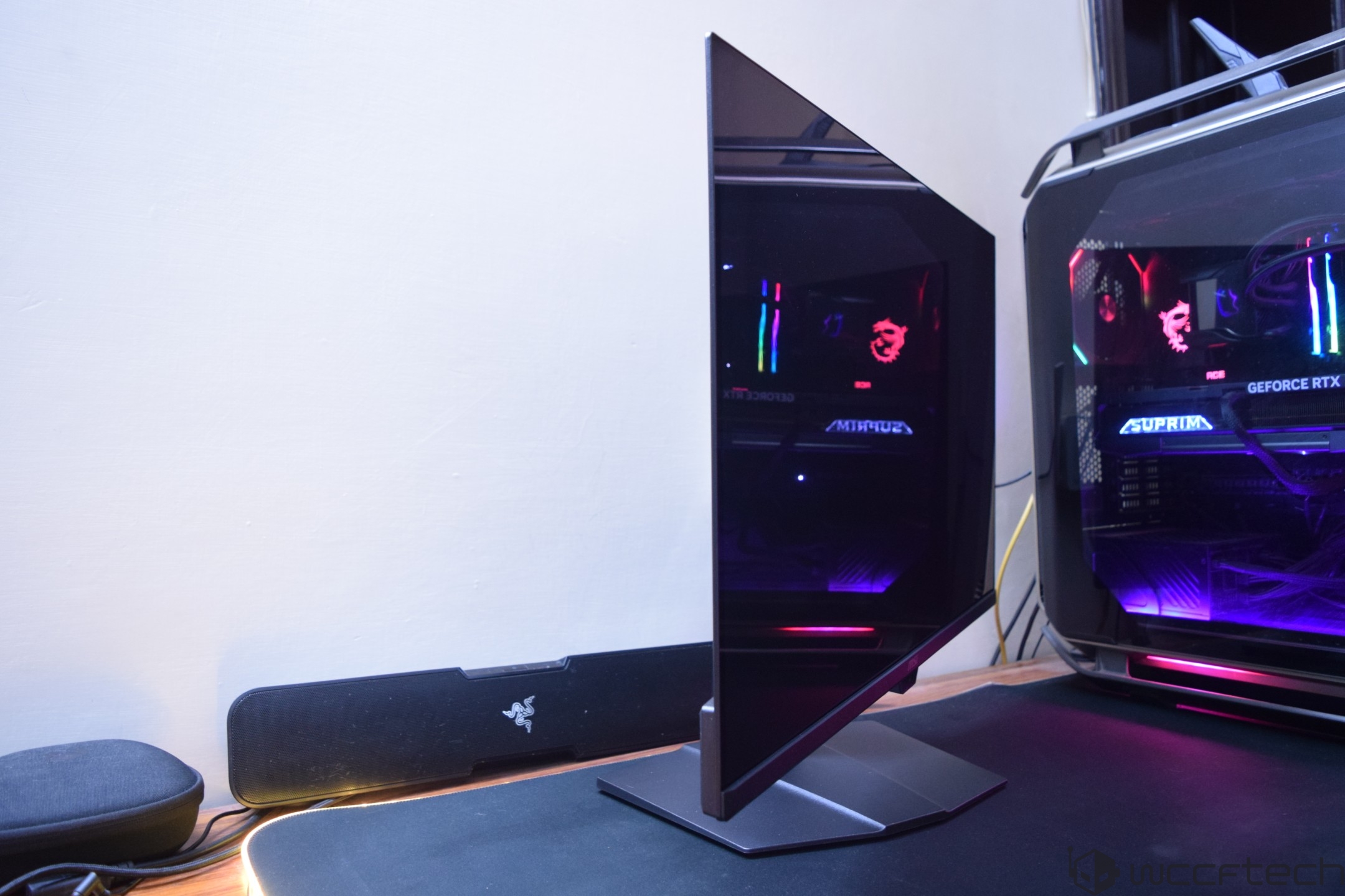 A gaming setup featuring a curved monitor and a desktop with a visible 'GEFORCE RTX' logo, illuminated in RGB, alongside a Razer soundbar on a desk.