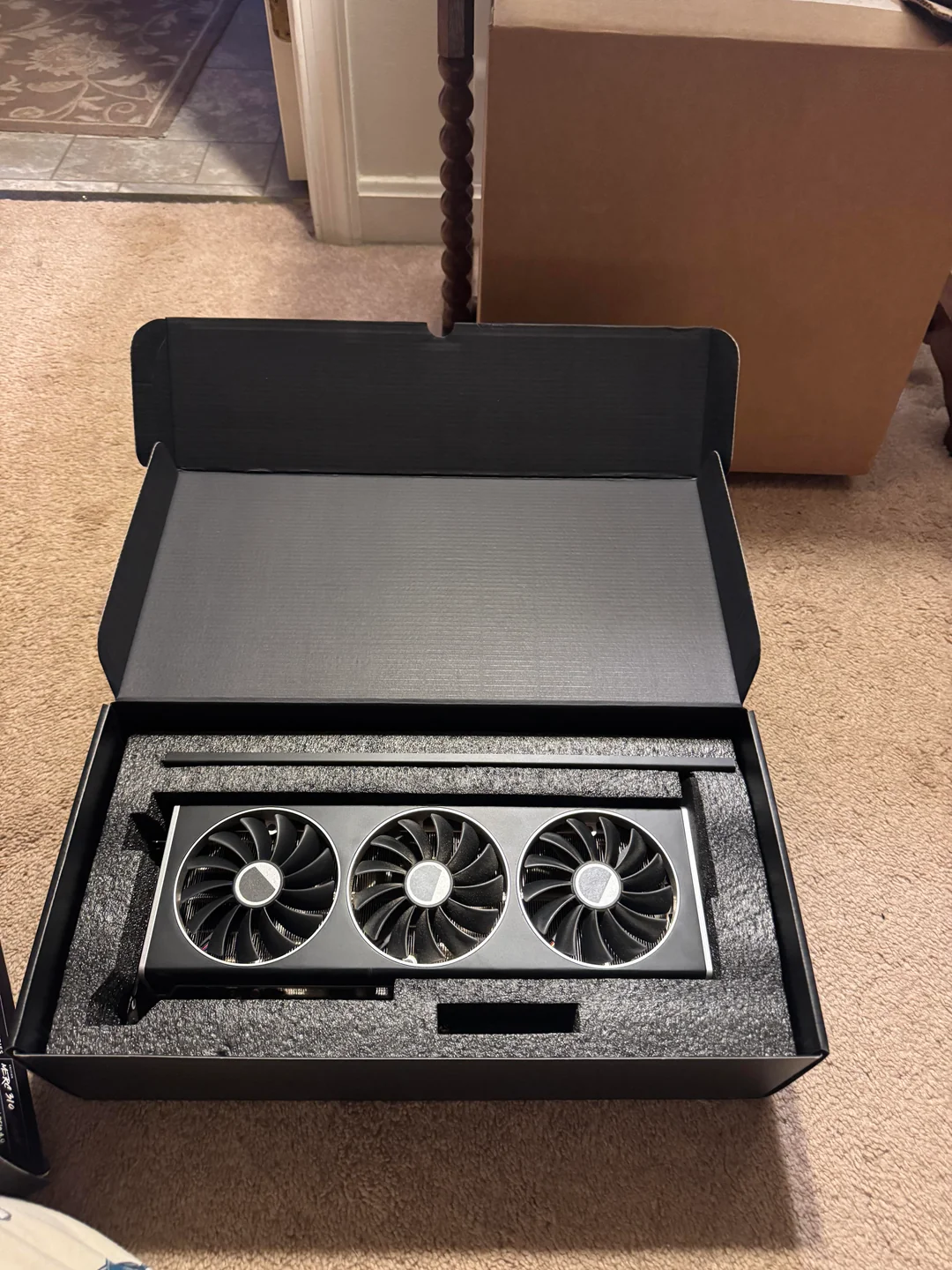 A graphics card with three cooling fans is displayed in an open black box on a carpeted floor.