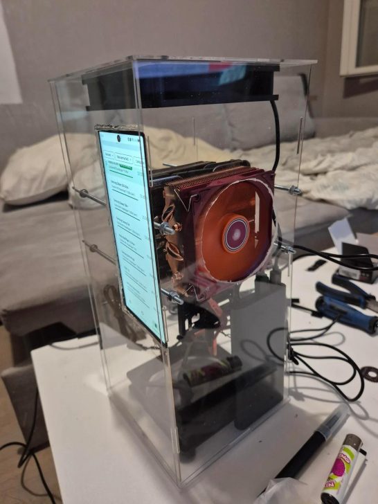 A smartphone is used as a display in a homemade computer encased in a transparent container, featuring a prominent red CPU cooler.